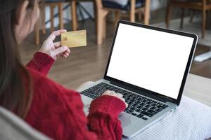 conectados compras. mulher comprando computador portátil usando crédito cartão sentado em sofá às casa foto