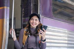 jovem ásia mulher dentro moderno trem estação fêmea mochileiro passageiro em pé usando telefone esperando para trem às trem estação para ir em feriado foto