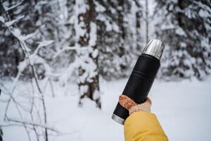 Preto fosco garrafa térmica dentro mão. viajante com luvas. beber caloroso chá e café a partir de uma garrafa térmica em uma andar dentro inverno. coberto de neve floresta em uma quieto dia. brilhante roupas foto