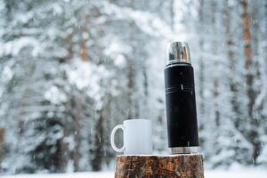 Preto fosco garrafa térmica e branco caneca com quente beber em toco. caminhada e piquenique funcionários. bebendo caloroso chá dentro a inverno geada. minimalista sujeito tiro foto