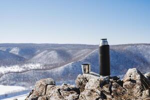 beber chá a partir de uma garrafa térmica dentro a montanhas. viagem através a rochoso colinas dentro a inverno temporada. uma ensolarado dia. uma Preto garrafa térmica com uma caneca contra a fundo do natureza. caminhada café da manhã às a topo foto