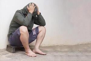 depressivo ásia homem desgasta moletom com capuz camisa, shorts, nu pés, senta magro velho parede em pavimento. conceito, vida problemas crise a partir de mau economia, pobreza. preocupado sobre desemprego. depressão. dor de cabeça. foto