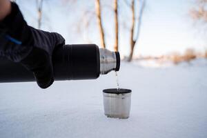 derramar quente chá a partir de uma garrafa térmica para dentro uma caneca. piquenique dentro natureza. inverno viagem. aço caneca com café. comendo em uma caminhada. metal pratos para turistas. foto