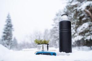 uma Preto fosco garrafa térmica, uma faca e uma caneca com uma abeto galho ficar de pé em uma coberto de neve coto.quente chá dentro a inverno geada.minimalista sujeito tiro foto