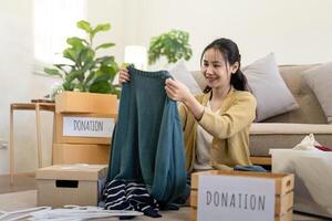mulher ásia segurando doação caixa cheio com roupas e selecionar roupas. conceito do doação e roupas reciclando. ajudando pobre pessoas foto