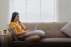 feliz mulher vestindo fones de ouvido assistindo e ouvindo vídeo em computador portátil e ouvindo música sentado em uma sofá às casa foto