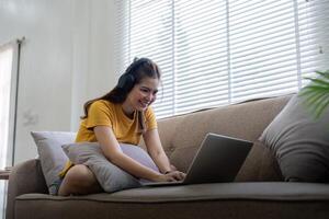 feliz mulher vestindo fones de ouvido assistindo e ouvindo vídeo em computador portátil e ouvindo música sentado em uma sofá às casa foto