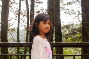 a ásia pequeno menina dentro grandes manga camisa em pé e posando em a sacada com natureza ou árvores fundo foto