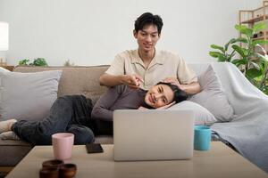 feliz casal ásia sentar relaxar em sofá dentro vivo quarto assistindo vídeo em computador portátil juntos às casa foto