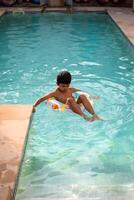 feliz indiano Garoto natação dentro uma piscina, criança vestindo natação traje ao longo com ar tubo durante quente verão Férias, crianças Garoto dentro grande natação piscina. foto