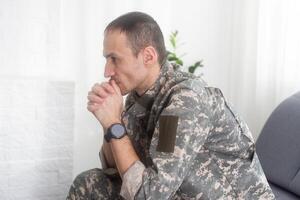 retrato do calma sério caucasiano militares homem vestindo camuflar uniforme e boné sentado em uma sofá, tendo depressivo facial expressão. foto