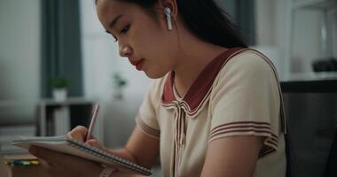 seletivo foco, lado visualizar, fêmea escritor vestem sem fio fones de ouvido sentado às escrivaninha enquanto escrevendo notas às lar, criativo pensamentos para diário, idéia e inspiração foto