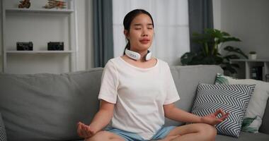 feliz jovem mulher vestem automático massagem pescoço meditando fazendo uma lótus pose às casa com olhos fechado, relaxante corpo e mente sozinho em sofá dentro a vivo quarto, relaxamento estilo de vida foto