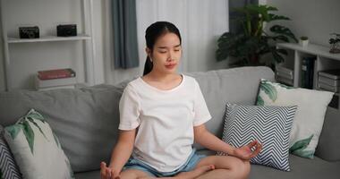 Alto ângulo visualizar, feliz jovem mulher vestem sem fio fones de ouvido meditando fazendo uma lótus pose às casa com olhos fechado, relaxante corpo e mente sozinho em sofá dentro a vivo quarto, relaxamento estilo de vida foto