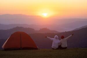 casal é sentado de a barraca durante durante a noite acampamento enquanto olhando às a lindo cênico pôr do sol sobre a montanha para ao ar livre aventura período de férias viagem conceito foto