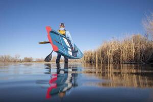 Senior masculino remador é lançamento uma ficar de pé acima paddleboard em uma calma lago dentro cedo primavera, rã perspectiva a partir de a açao Câmera às água nível foto