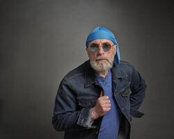 retrato do Senior, confiante, bonito homem com cinzento barba dentro crânio boné e caminhoneiro Jaqueta vestindo volta azul oculos de sol foto