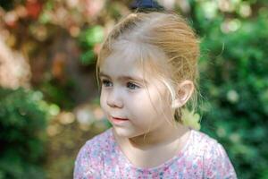 adorável pequeno menina com Loiras cabelo olhando longe dentro lindo jardim configuração foto