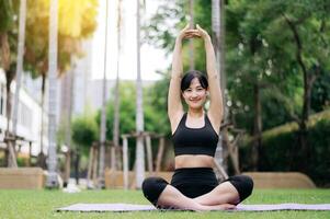 retrato em forma jovem ásia mulher 30s vestindo Preto roupa de esporte ouço relaxante música enquanto alongamento músculo antes ioga exercício em ioga esteira dentro verde natureza parque. bem estar conceito. foto