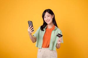 a conveniência do Móvel compras com a encantador jovem ásia mulher dentro dela 30s, vestindo laranja camisa e verde saltador, usando Smartphone enquanto segurando crédito cartão em amarelo estúdio fundo. foto