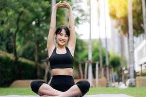 retrato em forma jovem ásia mulher 30s vestindo Preto roupa de esporte ouço relaxante música enquanto alongamento músculo antes ioga exercício em ioga esteira dentro verde natureza parque. bem estar conceito. foto