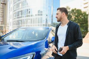 retrato do uma jovem homem com uma Móvel telefone dentro dele mão perto uma carro em uma verão rua. foto