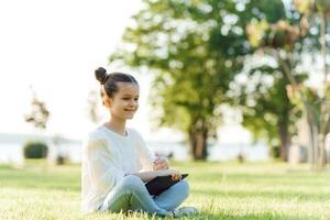 pequeno menina sentado em Relva e jogando tábua pc, tonificação foto. foto