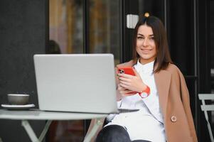 jovem feliz o negócio senhora parece às Smartphone tela com cheio de dentes sorrir enquanto sentado lado de fora em terraço com computador portátil e café, trabalhando remotamente conectados verificação relatórios e obtendo pronto para encontro foto