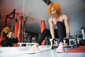 jovem desportivo mulher amarrar sapatos enquanto sentado em chão dentro academia. foto