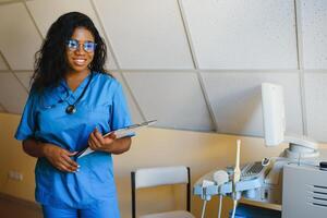 jovem afro americano fêmea médico em pé dentro clínica escritório perto moderno ultrassom máquina. foto