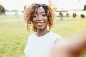 imagem do uma lindo jovem africano feliz mulher caminhando ao ar livre dentro uma Primavera parque levar uma selfie de Câmera faço Paz gesto. foto