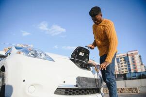 fechar acima do africano americano homem conectando cobrando cabo para elétrico carro. jovem masculino em pé perto dele moderno auto com couro mala de viagem dentro mão. foto