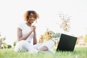 jovem sorridente Preto mulher sentado ao ar livre em Relva com computador portátil, digitando, surfar Internet, tendo café. tecnologia, comunicação, Educação e controlo remoto trabalhando conceito, cópia de espaço foto