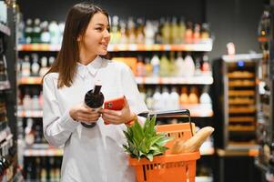 compras mulher olhando às a prateleiras dentro a supermercado. retrato do uma jovem menina dentro uma mercado loja com compras carrinho às álcool setor. foto