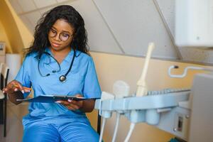 jovem alegre africano mulher operador do a ultrassom digitalização máquina analisando diagnóstico resultados do paciente. jovem sorridente africano médico trabalhando em uma moderno ultrassom equipamento. foto