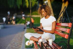 jovem lindo brasileiro fêmea com Preto encaracolado afro cabelo com tábua, enquanto sentado ao ar livre em a de madeira Banco dentro uma parque. foto