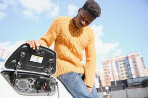 elétrico carros, ev conceito, eco amigáveis combustível. retrato do jovem sorridente Preto homem, recarregar dele moderno luxo elétrico carro foto