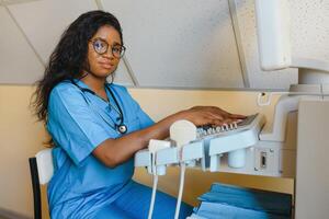 sorridente africano mulher médico com ultrassom scanner dentro mão, trabalhando em moderno ultrassom digitalização máquina dentro luz quarto dentro clínica. retrato do 4d ultrassom digitalização máquina operador foto