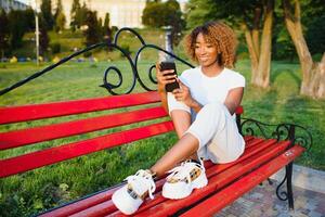 retrato do jovem africano americano mulher sentado em parque Banco com celular dentro mão foto