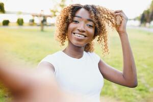 imagem do uma lindo jovem africano feliz mulher caminhando ao ar livre dentro uma Primavera parque levar uma selfie de Câmera faço Paz gesto. foto