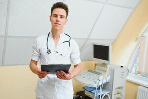 encantador jovem masculino médico , sentado às dele escritório perto ultrassom digitalização máquina. bonito amigáveis médico desfrutando trabalhando às a hospital, cópia de espaço. ultrassônico conceito foto