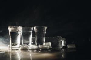 fechar-se Visão do garrafa do vodka com óculos em pé em gelo em Preto. foto