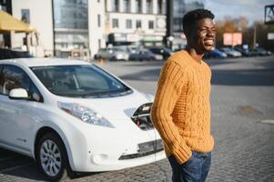 urbano, elétrico veículo, eco conceito. jovem Preto esfolado homem, esperando para dele elétrico carro cobrando foto