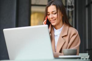 jovem trabalhador autonomo mulher desfrutando café enquanto trabalhando ao ar livre em uma computador portátil computador conectado para público Wi-fi. à moda aluna menina estudando conectados dentro cidade. foto