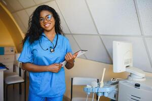 jovem afro americano fêmea médico em pé dentro clínica escritório perto moderno ultrassom máquina. foto