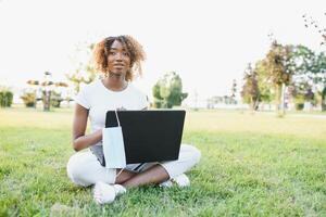 humano e tecnologia conceito. encantador africano mulher com curto duende Penteado desfrutando ensolarado clima, sentado em a gramado dentro frente do computador portátil computador dentro a público jardim, esperando para dela amigo foto