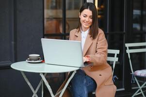 jovem trabalhador autonomo mulher desfrutando café enquanto trabalhando ao ar livre em uma computador portátil computador conectado para público Wi-fi. à moda aluna menina estudando conectados dentro cidade. foto