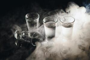 fechar-se Visão do garrafa do vodka com óculos em pé em gelo em Preto. foto