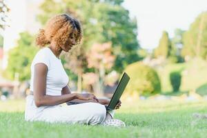 humano e tecnologia conceito. encantador africano mulher com curto duende Penteado desfrutando ensolarado clima, sentado em a gramado dentro frente do computador portátil computador dentro a público jardim, esperando para dela amigo foto