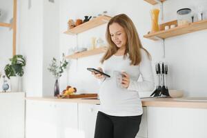 cenário do lindo casualmente vestido jovem morena fêmea esperando bebê, tendo manhã chá dentro moderno à moda cozinha, sorridente. antecipação, expectativa, gravidez e maternidade conceito foto